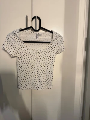 Vit blommig topp från Hollister - Söt vit topp från Hollister med små svarta blommor över hela tyget. Modellen är croppad med korta ärmar och har en rundad, djup urringning framtill samt små dekorativa knappar. Perfekt för dig som gillar en feminin och trendig stil.