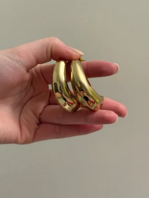 Chunky guldiga hoops örhängen - Säljer ett par chunky, breda hoops i guldtonad metall. Örhängena har en blank, spegelblank finish och en rundad, tjock form som ger ett statement-intryck. Perfekta för dig som gillar trendiga accessoarer med attityd.
