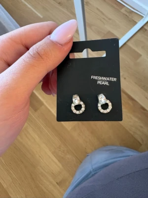 Silverörhängen med sötvattenspärlor - Snygga örhängen i silverfärg med små gnistrande stenar i en cirkelform och en sötvattenspärla upptill. Perfekta för dig som gillar stilrena men ändå unika accessoarer. Kommer på originalkort.