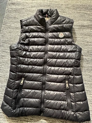 Svart dunväst från Moncler - Snygg svart dunväst från Moncler med quiltad design och hög krage. Västen har två dragkedjeförsedda fickor framtill och en diskret Moncler-logga på bröstet. Tillverkad i glansigt material som ger en exklusiv känsla.