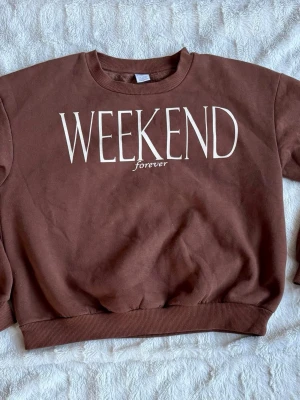 Brun sweatshirt med WEEKEND-tryck - Cool brun sweatshirt med texten 'WEEKEND forever' i vitt framtill. Tröjan har rund hals, ribbade muddar vid ärmslut och nederkant samt en avslappnad passform. Perfekt för chill dagar och enkel att matcha med jeans eller mjukisbyxor.