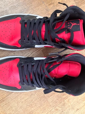 Air Jordan 1 Mid röd/svart/vit - Säljer ett par Air Jordan 1 Mid sneakers i rött, svart och vitt. Skorna har klassisk hög siluett, svart snörning och ikoniska Jumpman-loggan på plösen. Ovandelen är i läder och färgblocken ger en retro vibe. Perfekta för dig som gillar streetwear och sneakerskultur.