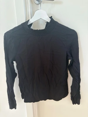 Svart blus med smockad krage - Stilren svart blus med långärmad design och smockad hög krage. Blusen har även smockade muddar vid ärmsluten och en lätt rynkad effekt i tyget. Perfekt till jeans eller kjol för en trendig look.