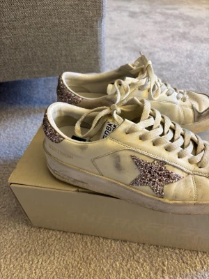 Golden Goose vita sneakers med glitterstjärna - Snygga vita sneakers från Golden Goose med glitterdetaljer i form av en stjärna på sidan och glitter på hälen. Skorna har vita snören, perforerad tå och beige mockadetaljer framtill. Tillverkade i läder och mocka, perfekta för en trendig streetstyle-look.
