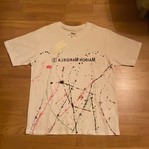 Vit t-shirt med färgstänk från Maison Margiela - Vit t-shirt från Maison Margiela med tryckt logga och färgstänk i flera färger. Storlek L. Prislappen är tyvärr inte längre kvar. Använd ett fåtal gånger.
