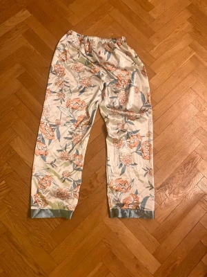 Blommiga satinpyjamasbyxor - Snygga pyjamasbyxor i glansigt satinmaterial med blommigt mönster i persika, grönt och blått på ljus botten. Byxorna har resår i midjan och ljusblå kant vid bensluten. Perfekta för chillkvällar hemma.Skriv gärna för mått! 💕