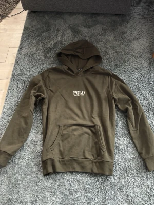 Olivgrön hoodie från Polo Ralph Lauren - Säljer en olivgrön hoodie från Polo Ralph Lauren med vit logotyp på bröstet. Hoodien har en stor huva, ribbade muddar och en klassisk magficka. Materialet är mjukt och bekvämt, perfekt för chill dagar. Passar dig som gillar stilren design och ikoniska märken.