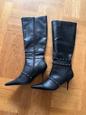 Svarta knähöga boots med nitar - Säljer ett par svarta knähöga boots med spetsig tå och smal klack. Skorna har dekorativa remmar med silverfärgade nitar runt ankeln och skaftet. Tillverkade i ett slätt, syntetiskt material med dragkedja på insidan för enkel påtagning.