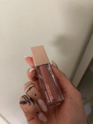 Maybelline Lifter Gloss i nude - Maybelline Lifter Gloss med hyaluronsyra i en snygg nude-rosa nyans. Kommer i en stilren, fyrkantig plastförpackning med beige lock och genomskinlig flaska som visar färgen. Perfekt för glansiga och återfuktade läppar.