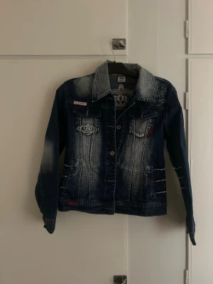 Mörkblå Y2K jeansjacka Blue Queen - Snygg mörkblå jeansjacka från Blue Queen med slitna detaljer. Jackan har två bröstfickor med lock, varav en är dekorerad med ett broderat kronemärke. Klassisk krage och knappstängning framtill.
