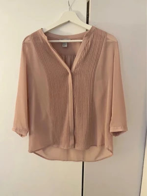 Ljusrosa blus med veckad front - Snygg ljusrosa blus med trekvartsärmar och v-ringning. Framsidan har stilrena veckade detaljer som ger en elegant touch. Tillverkad i ett tunt, lätt och något transparent material som faller fint. Perfekt att matcha med jeans eller kjol.