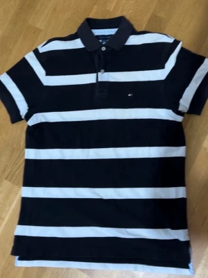 Randig pikétröja från Tommy Hilfiger - Säljer en svart och vit randig pikétröja från Tommy Hilfiger. Tröjan har klassisk krage, korta ärmar och knappar vid halsen. Har en liten logga på bröstet. Perfekt för en avslappnad och stilren look. Storlek S-M