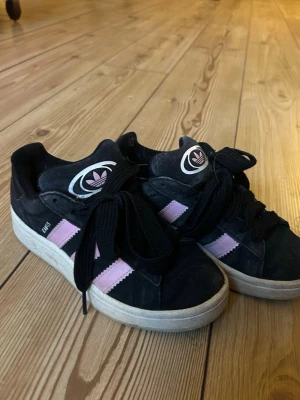 Adidas svart/lila sneakers - Snygga Adidas sneakers i svart mocka med lila ränder på sidorna och vit sula. Klassisk Adidas-logga på plösen och svarta skosnören. Perfekta för dig som gillar streetstyle och vill ha något unikt på fötterna.