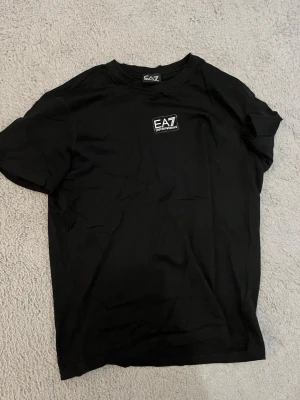 Svart EA7 t-shirt i bomull - Snygg svart t-shirt från EA7 Emporio Armani med liten logga på bröstet. Klassisk rund hals och korta ärmar. Tillverkad i mjuk bomull som känns skön mot huden. Perfekt till jeans eller shorts för en clean och stilren look.