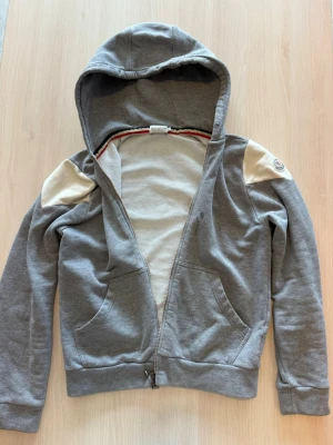 Grå hoodie med dragkedja från Moncler - Snygg grå hoodie från Moncler med dragkedja framtill och två fickor. Hoodien har vita detaljer på axlarna och Moncler-logga på vänster ärm. Tillverkad i mjuk bomullsblandning och har en klassisk huva. Perfekt för en avslappnad och trendig look. Priset går att diskuteras 