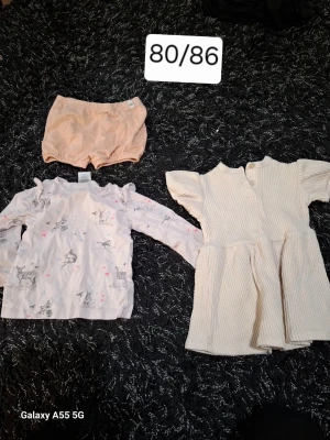 Barnklädespaket 80/86 ljusa toner - Säljer ett paket med tre plagg i storlek 80/86: ett par ljusrosa shorts, en långärmad vit tröja med djurmotiv och volangdetaljer samt en cremevit ribbad klänning med kort ärm och knappar bak. Perfekt för dig som gillar mjuka färger och söta detaljer.
