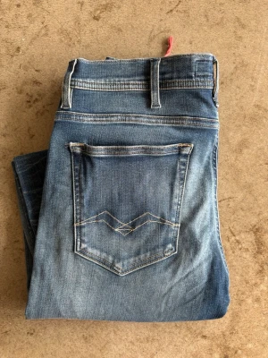 Blå jeans från Replay - Säljer ett par klassiska blå jeans från Replay jeans med snyggt slitna detaljer och kontrastsömmar. Jeansen har en rak passform, fem fickor och en cool broderad bakficka. Perfekta för en avslappnad och trendig look.