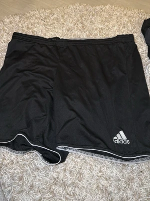 Svarta sportshorts från Adidas - Svarta sportshorts från Adidas med vit logga på vänster ben och vita detaljer längs kanterna. Tillverkade i ett lätt och ventilerande syntetmaterial, perfekta för träning eller sportaktiviteter. Klassisk midja och avslappnad passform.