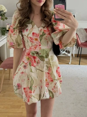 Blommig kortklänning med puffärm - Supersöt kortklänning med blommigt mönster i ljusa toner, v-ringad hals och puffiga korta ärmar. Klänningen har en markerad midja och volangig kjol som ger en romantisk vibe. Perfekt för sommarens alla tillfällen!