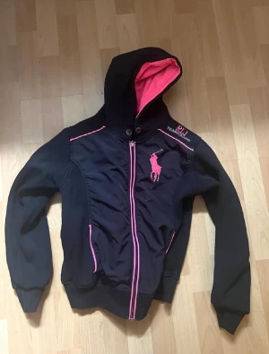 Svart hoodiejacka RL1 Teamracing - Svart hoodiejacka från Ralph lauren med rosa detaljer och broderad logga på bröstet. Jackan har dragkedja framtill, huva med rosa insida och långa ärmar. Materialet är troligtvis polyester eller bomullsmix, perfekt för dig som gillar sportig stil. En fläck i luvan som jag ej testat att få bort. 