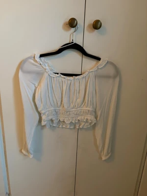 Vit off shoulder crop top med volanger - Säljer en söt vit off shoulder crop top med långa ärmar och volangdetaljer vid halsringning och nederkant. Perfekt att matcha med höga jeans eller kjol. Aldrig använd men saknar lapp.