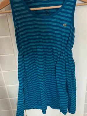 Lacoste randigt linne XS 36 turkos blå krokodil topp sommar - Snygg blå kortklänning från Lacoste med horisontella ränder i två blå nyanser. Klänningen är ärmlös, har rund halsringning och ett litet Lacoste-märke på bröstet. Tillverkad i mjuk bomull, perfekt för varma dagar.