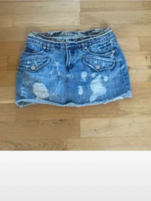 Vintage jeans kjol - Säljer en kort blå jeanskjol med slitna detaljer och rå kant. Kjolen har två fickor fram med lock och knappar samt två bakfickor. Perfekt för dig som gillar en avslappnad och trendig stil.