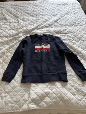 Mörkblå sweatshirt Tommy Hilfiger - Mörkblå sweatshirt från Tommy Hilfiger med klassisk logga och röd-vit-blå detalj på bröstet. Tröjan har rund halsringning, ribbade muddar och är tillverkad i mjuk bomullsblandning. Perfekt för en avslappnad och stilren look. Det är storlek 152 barn storlek