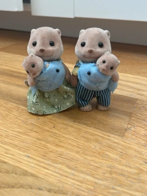Sylvanian Families bäverfamilj - Två söta bäverfigurer från Sylvanian Families med babybävrar i blå bärselar. Ena vuxna har grön klänning med blommönster, den andra har blåvitrandiga byxor och gul skjorta. Figurerna är gjorda av mjuk plast och tyg.
