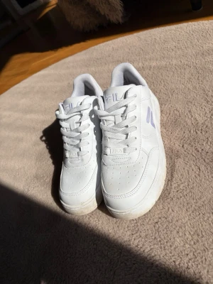 Vita Fila sneakers med lila detaljer - Snygga vita sneakers från Fila med klassisk låg profil och lila logga på sidan. Skorna har perforerad tå för ventilation, vadderad plös och krage samt vita skosnören. Tillverkade i syntetmaterial och passar perfekt till streetwear-stilen.