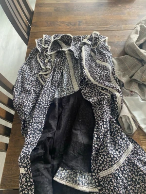 Blommig volangklänning med spetsdetaljer - Supersöt blommig klänning med volanger och spetsdetaljer. Klänningen har mörk botten med små vita blommor, långa ärmar och markerad midja. Perfekt för dig som gillar romantisk och trendig stil.