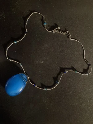 Halsband med blått hänge och pärlor - Unikt halsband med ett stort blått ovalt hänge i glas och små blå pärlor längs den tunna silverfärgade kedjan. Perfekt för dig som vill sticka ut med en färgklick och cool design. Justerbart spänne för bästa passform.OBS! Materialet är okänt, kan ej garantera att det är rostfritt.