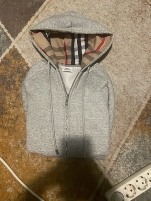Grå hoodie med Burberry-mönstrad huva - Snygg grå zip-hoodie från Burberry med klassiskt rutmönster i huvan. Dragkedja framtill och justerbara snören i huvan. Perfekt för dig som gillar stilrena plagg med en twist. Tillverkad i mjukt material för extra komfort.