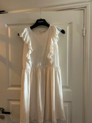 Vit volangklänning från Kappahl - Söt vit klänning från Kappahl med volangdetaljer längs axlarna och broderade hålmönster. Klänningen har rund halsringning och är tillverkad i ett lätt material, perfekt för varma dagar.