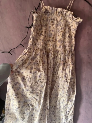 Ljusblommig långklänning med volang - Söt långklänning med smala axelband och ribbat stretch material vid bysten. Klänningen är ljus med lila blommigt mönster och har smock upptill för en bekväm passform. Perfekt för varma dagar och somriga tillfällen.