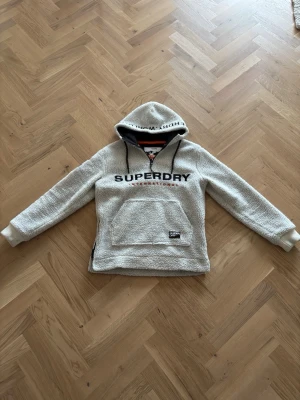 Superdry vit teddy hoodie - Mysig vit teddy hoodie från Superdry med svart logga broderad över bröstet och detaljer på ärmen. Hoodien har stor känguruficka framtill, dragsko i huvan och ribbade muddar. Perfekt för dig som gillar streetwear och vill ha något riktigt bekvämt och snyggt.