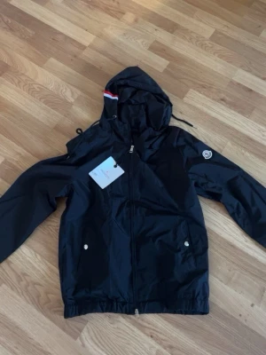 Svart vindjacka från Moncler - Snygg svart vindjacka från Moncler med huva och dragkedja framtill. Jackan har två fickor med tryckknappar och en Moncler-logga på ärmen. Huvan har en rödvit rand och dragsko för justering. Tillverkad i lätt, vattenavvisande material.