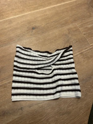 Randig stickad bandeau tubtopp - Säljer en stickad bandeau tubtopp med svart och vit randigt mönster. Toppen har en twistad detalj framtill och är ribbad för extrautdelning stretch och passform. Perfekt för dig som vill ha en enkel men snygg topp till sommaren.