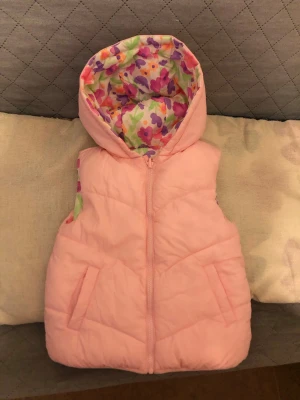 Vändbar blommig rosa dunväst - Supersöt vändbar dunväst med ena sidan i ljusrosa och andra sidan med färgglatt blommönster i lila, rosa, orange och grönt. Västen har huva och dragkedja framtill samt två fickor. Perfekt för dig som vill ha två looks i en och gillar färg och mönster.