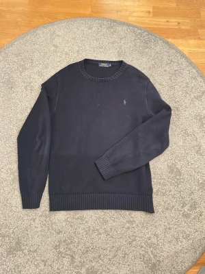 Mörkblå stickad tröja Polo Ralph Lauren - Mörkblå stickad tröja från Polo Ralph Lauren med rund halsringning och ribbade muddar. Klassisk grön broderad logga på bröstet!