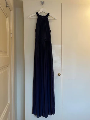 Marinblå långklänning från Moments i storlek 34 - Elegant marinblå långklänning med halterneck och knyt i nacken. Klänningen har ett plisserat, flytande tyg och markerad midja som ger en snygg siluett. Perfekt för bal eller festligare tillfällen!