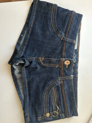 Mörkblå jeansshorts med dragkedja - Säljer ett par mörkblå jeansshorts från Lee med snygga kontrastsömmar och detaljer i brunt. Shortsen har klassiska fickor fram och bak, samt en liten dragkedjedetalj framtill. Perfekta för sommaren och riktigt sköna att bära. Låg midja och midjemåttet är 41☺️