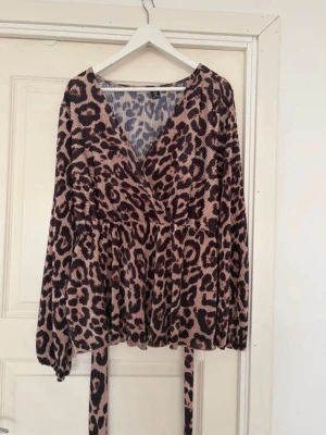Leopardmönstrad omlottopp med volang 3XL - Säljer en leopardmönstrad omlottopp med v-ringning och långa, lösa ärmar. Toppen har volangdetaljer och knytband i midjan för en snygg siluett. Perfekt för dig som vill sticka ut med ett djurmönster!