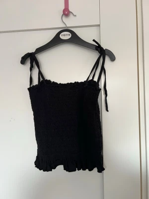 Svart smockad linnetopp med knytband - Supersöt svart linnetopp med smockad struktur och volangkant nertill. Axelbanden knyts på axlarna för en justerbar passform. Perfekt till jeans eller kjol för en trendig look.