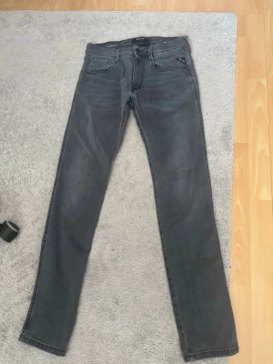 Svarta slim jeans från Replay - Säljer ett par svarta slim fit jeans från Replay med klassisk femficksdesign och diskreta slitningar. Jeansen har raka ben och är tillverkade i mjukt denimtyg som sitter snyggt. Perfekta för dig som gillar en stilren och enkel look.