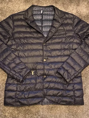 Blå dunväst från Strömberg  - Stilren svart dunväst med kavajkrage och knäppning framtill. Västen har två framfickor, quiltad design och lätt glansigt material. Perfekt för dig som vill ha en modern twist på klassisk dunväst. Hör av er vid frågor 