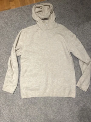 Beige hoodie i mjukt material - Säljer en stilren beige hoodie med luva och långa ärmar. Hoodien är tillverkad i ett mjukt, stickat material och har ribbade muddar vid ärmslut och nederkant. Perfekt för en avslappnad och clean look.