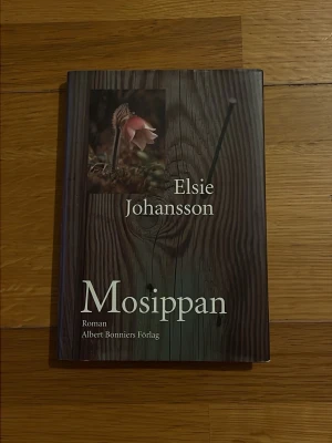 Mosippan av Elsie Johansson - Romanen 'Mosippan' av Elsie Johansson har ett omslag med träliknande mönster och en bild på en rosa blomma. Boken är inbunden och utgiven av Albert Bonniers Förlag. Perfekt för dig som gillar svensk skönlitteratur och starka berättelser.