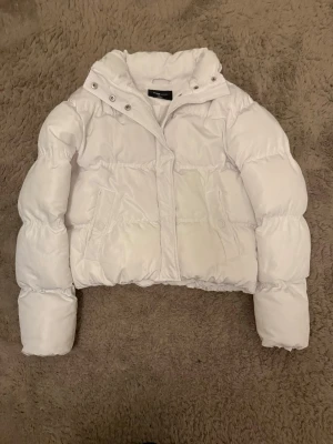 Vit kort pufferjacka med huva - Säljer en snygg vit pufferjacka med kortare passform och huva. Jackan har två sidofickor, dragkedja och tryckknappar framtill. Perfekt för kalla vinterdagar och ger en trendig look. Fodrad för extra värme och har en stilren design.