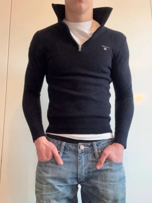 Mörkblå stickad tröja från GANT - Mörkblå stickad tröja från GANT med halv dragkedja och hög krage. Storleken är S men passar mer XS .Tröjan har lång ärm och diskret GANT-logga broderad på bröstet. Perfekt för lager på lager och ger en clean, klassisk look.